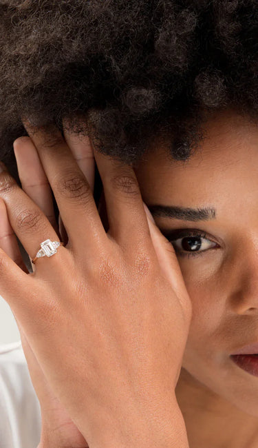 Woman's hand with "The Chapters" engagement ring, featuring an emerald cut diamond and gold band.