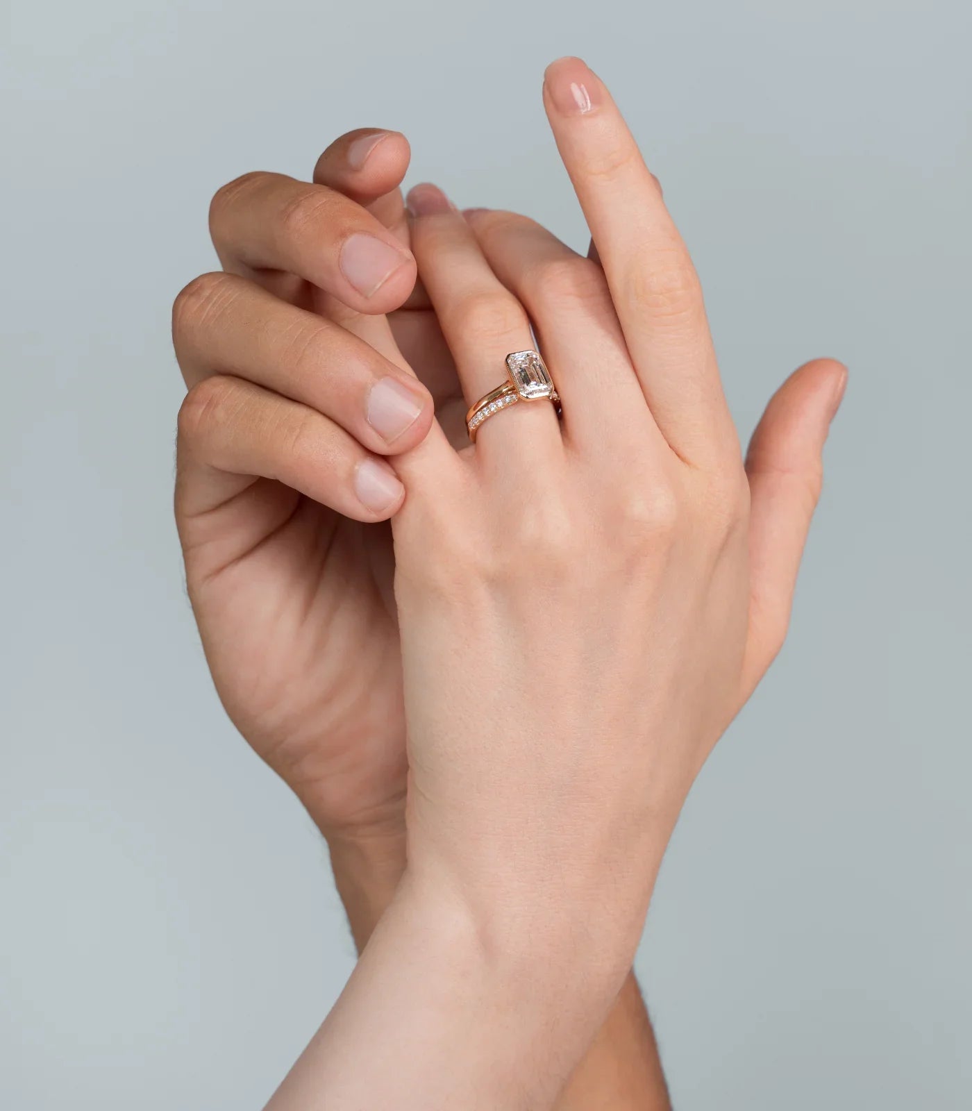 Close-up of "Keanis" bridal set. Emerald cut engagement ring with diamond band on a woman's hand, held by a man's hand.
