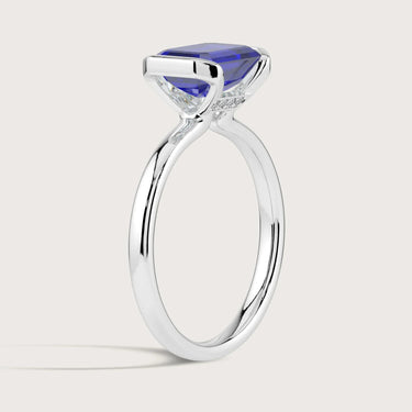 Lab-grown sapphire ring. Emerald cut blue gemstone in a 18k white gold solitaire half-bezel setting. Sparkly diamond accents.