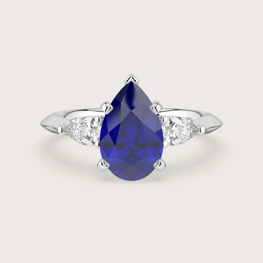 Lab-grown pear sapphire trilogy engagement ring. 18k white gold band, blue gemstone center, white side stones.